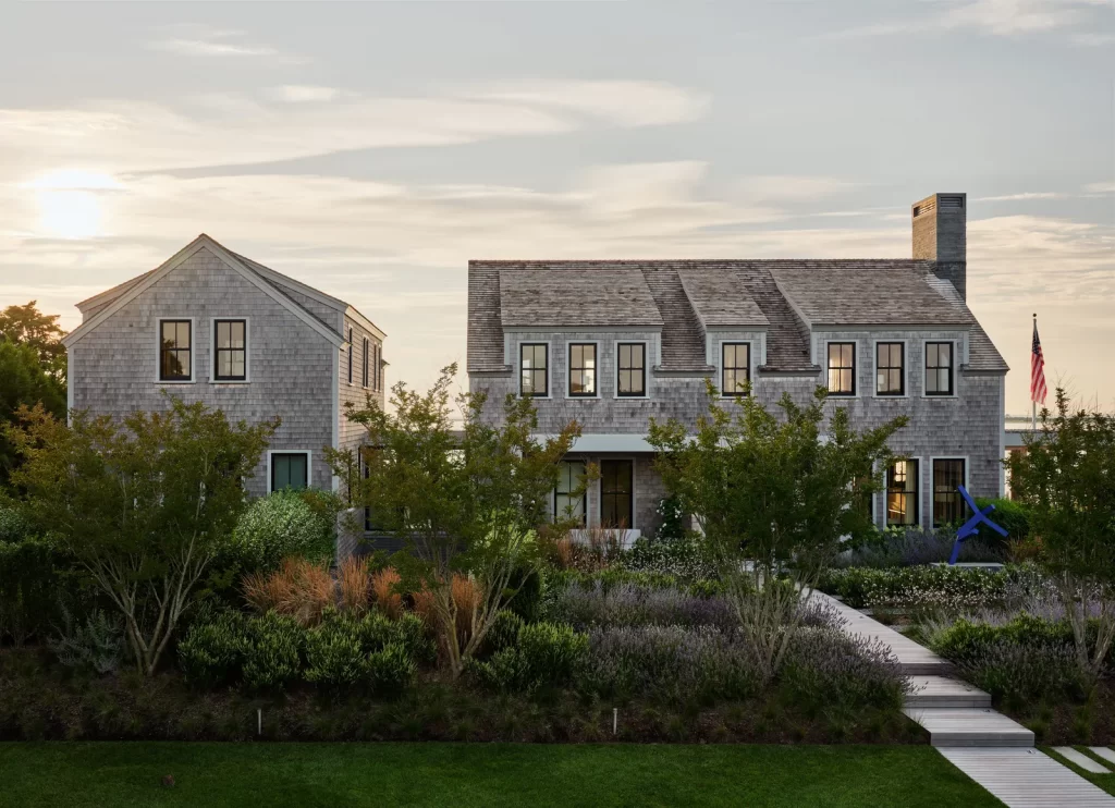 A large shingle-style coastal home with traditional New England architectural elements features symmetrical gabled roofs, double-hung black-framed windows, and a prominent central chimney, flanked by a detached guest house on the left, both structures clad in weathered gray cedar shingles, surrounded by meticulously landscaped gardens with native shrubs, ornamental grasses, and lavender, while a narrow wooden boardwalk leads through the lush greenery to the front porch, set against a calm sunset sky that bathes the entire scene in a warm, golden light, and an American flag waves in the background near the water's edge.