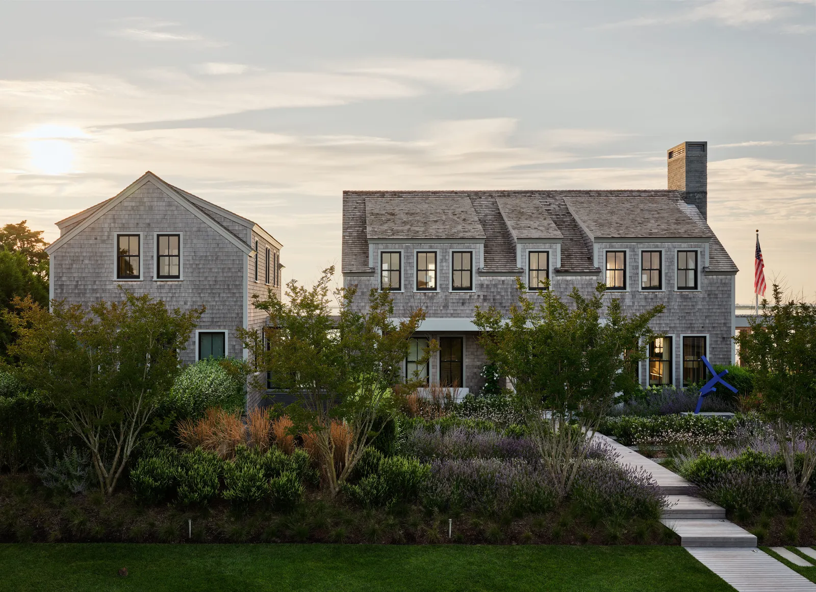 A large shingle-style coastal home with traditional New England architectural elements features symmetrical gabled roofs, double-hung black-framed windows, and a prominent central chimney, flanked by a detached guest house on the left, both structures clad in weathered gray cedar shingles, surrounded by meticulously landscaped gardens with native shrubs, ornamental grasses, and lavender, while a narrow wooden boardwalk leads through the lush greenery to the front porch, set against a calm sunset sky that bathes the entire scene in a warm, golden light, and an American flag waves in the background near the water's edge.