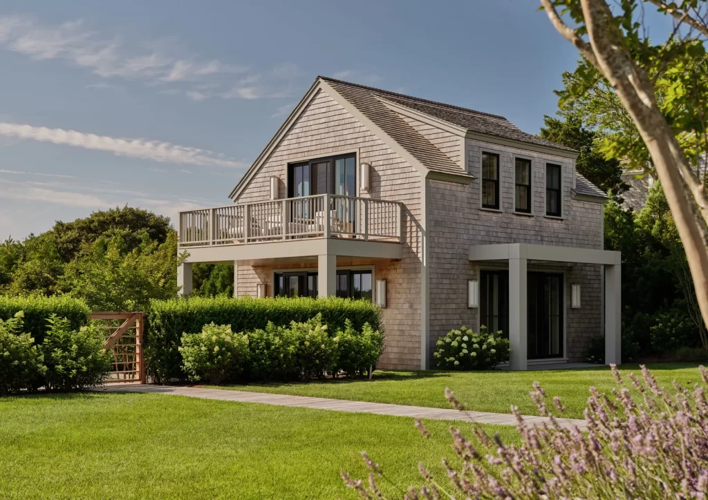 A charming two-story guest house with weathered cedar shingles, a pitched roof, and large black-framed windows sits amid lush manicured landscaping, featuring an upper-level balcony with a light-colored wooden railing, a covered ground-floor patio area supported by square columns, neatly trimmed hedges surrounding the structure, a wooden gate integrated into the greenery, and a stone walkway leading through a vibrant lawn bordered by flowering plants under a bright blue sky.