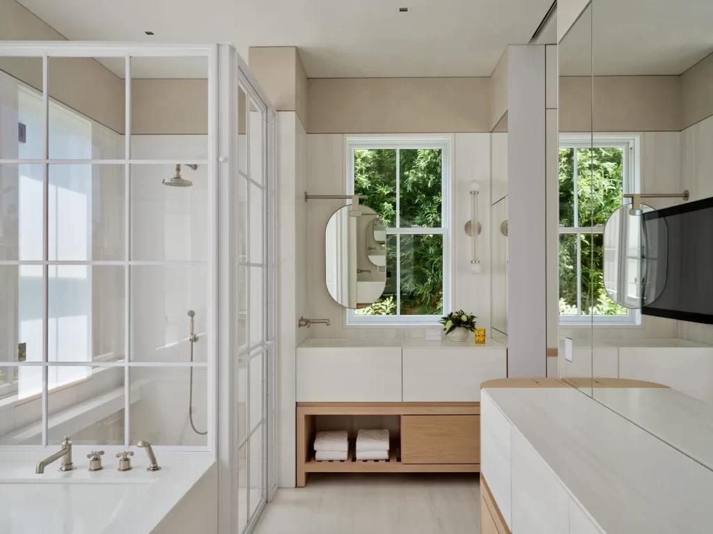 A bright, contemporary bathroom featuring a large soaking tub with polished chrome fixtures beside a walk-in shower enclosed in a grid-framed glass partition, dual vanity areas with minimalist white sinks and light wood cabinetry below, round mirrors set against vertical rectangular windows that frame lush green foliage outside, and a sleek mirrored wall reflecting the clean lines and calming palette of the space.