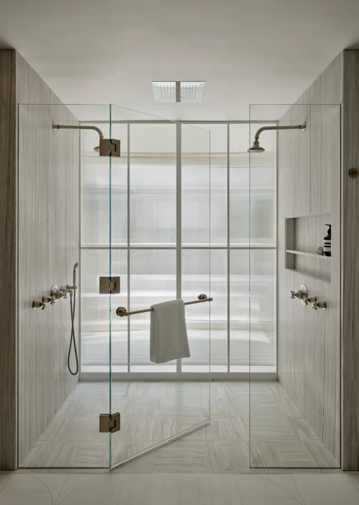 A luxurious walk-in shower enclosed by seamless glass panels with sleek brass hardware, featuring dual wall-mounted showerheads and a central rain shower above, all set against vertically textured stone walls and light-toned tiled flooring, with a minimalist towel bar holding a white towel on the door, built-in shelving for toiletries on the right wall, and soft diffused light filtering through a translucent glass-paneled window wall that adds privacy while maintaining a bright and airy atmosphere.