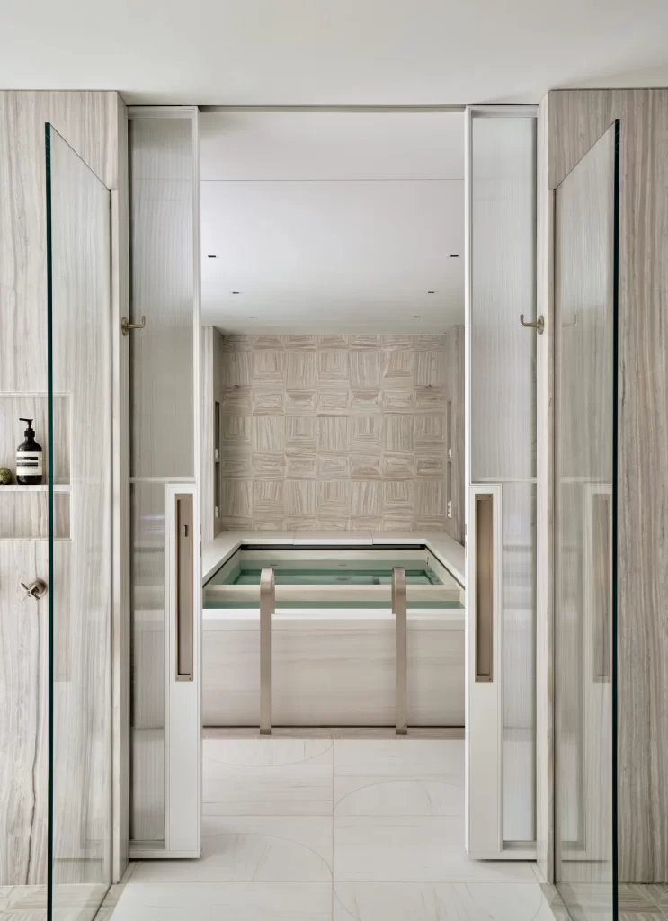 A serene spa-like room with a recessed plunge pool framed in soft-toned marble and brushed metal railings, surrounded by textured vertical stone walls and a geometric patterned feature wall at the far end, accessed through partially opened frosted glass sliding doors with sleek handles, all set beneath a clean white ceiling dotted with recessed lights that enhance the tranquil and minimalistic ambiance of the space.