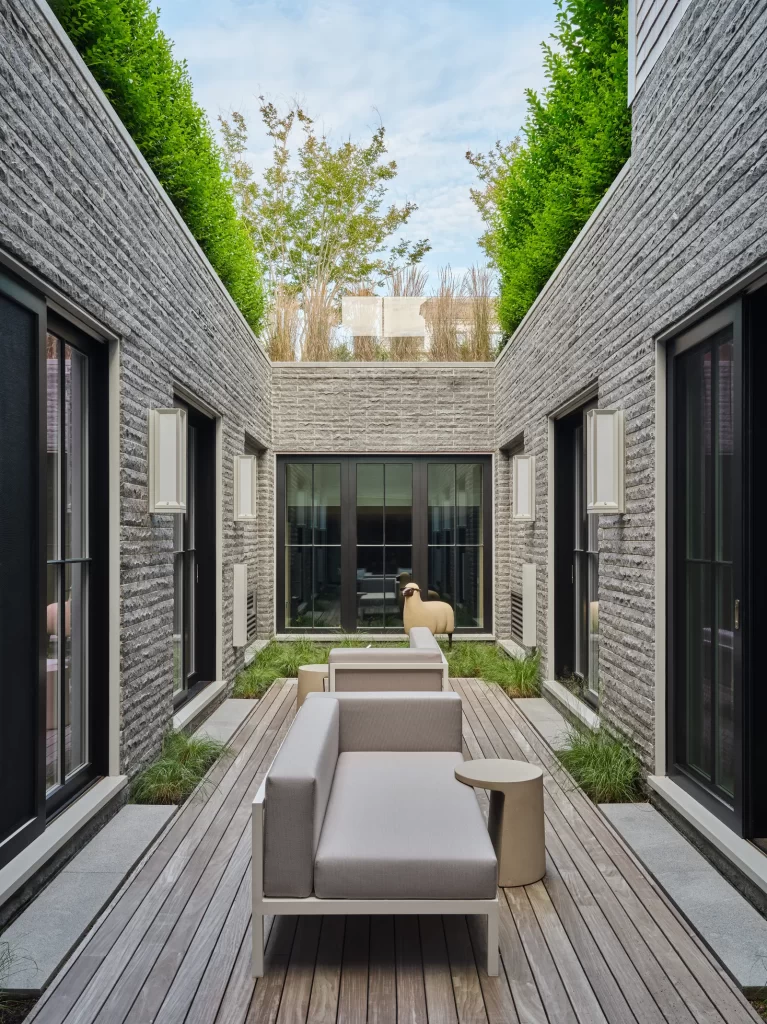 A narrow, modern courtyard framed by textured gray stone walls and large black-framed windows features minimalist outdoor furniture including pale taupe sofas and small cylindrical side tables placed on a wooden deck walkway flanked by patches of ornamental grass, while tall manicured hedges line the tops of the surrounding walls creating a lush green contrast against the structured architecture, and a whimsical sculptural sheep figure stands at the far end near a set of double glass doors, all beneath a soft blue sky with scattered clouds and framed by distant trees visible above the courtyard walls.