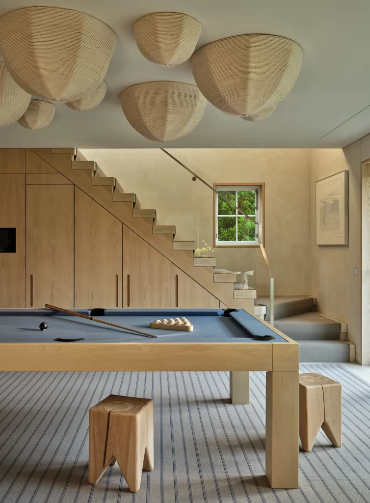A serene and stylish interior recreational room featuring a natural wood pool table with a navy blue felt top, billiard balls arranged in a triangle and cue sticks placed diagonally across the surface, set atop a subtly striped carpet, flanked by uniquely sculptural wooden stools, all framed by a staircase with integrated wooden cabinetry underneath and a minimalist glass railing, with a soft neutral-toned plaster wall finish, a square window providing a glimpse of green foliage outside, a framed artwork on the wall, and an arrangement of woven basket-style light fixtures dramatically adorning the ceiling.