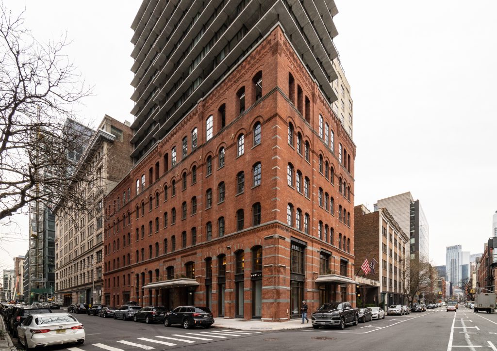 A striking red-brick corner building with arched windows and industrial architectural detailing sits prominently on a city intersection, its historical facade blending with modern urban life as it supports a contemporary residential tower that juts upward with cantilevered balconies, while pedestrians and parked cars line both streets under a gray overcast sky, flanked by a diverse mix of adjacent buildings including glass-clad structures and older masonry edifices, creating a vivid contrast between old and new in a bustling metropolitan environment.