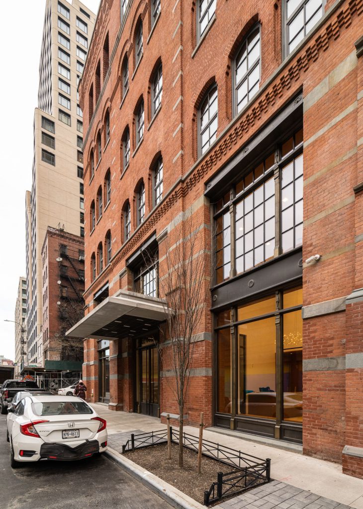 This image showcases a multi-story brick building with arched windows and industrial-style black metal window frames, blending historical architecture with modern upgrades. The ground floor features large storefront or lobby windows, and a canopy projects above the main entrance. Decorative gray stone banding accents the red brick façade, contributing to a refined historic character often found in renovated urban warehouses or loft-style buildings. A white car is parked at the curb, adding scale and context to the streetscape.