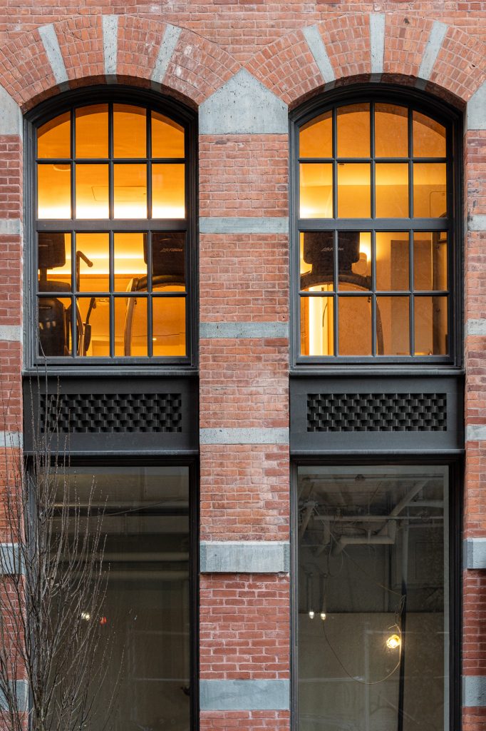 A symmetrical façade of a red brick building featuring two vertically aligned sets of large windows with black grilles and mullions, the upper arched windows glowing with warm interior light revealing gym equipment behind them, while the lower rectangular windows are unlit and partially obscured, all framed by alternating horizontal concrete bands and arched concrete keystones that emphasize the historic industrial architectural style.
