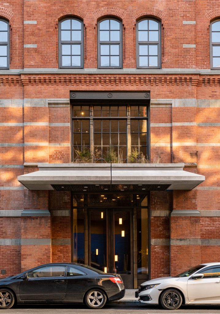 Centrally focused entrance of a historic red-brick building adorned with horizontally laid stone banding, featuring a large modern glass doorway flanked by tall bronze-toned metal frames beneath a prominent overhanging concrete canopy, topped by a window box planter with dry ornamental grasses and shrubs, while a tall industrial-style window above is divided by vertical iron mullions and framed in dark metal with decorative roundels, all illuminated by warm interior lighting and dappled sunlight casting golden reflections across the façade, with two parked cars grounding the scene in a contemporary urban context.