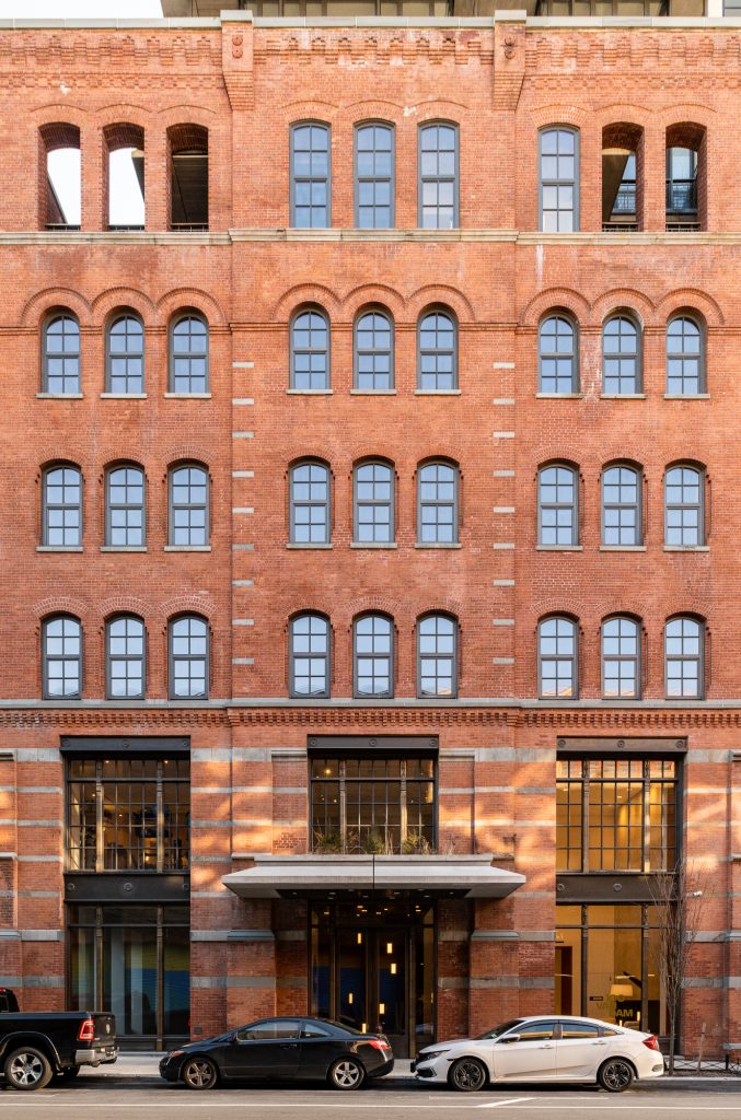 Symmetrically framed red-brick building facade featuring repetitive arched windows with dark mullions, flanked by strong vertical and horizontal stone banding elements that accentuate its historic industrial aesthetic, while the lower level presents modernized tall black-framed glass entryways and storefronts, including a central entrance with a concrete canopy above, where a row of parked vehicles lines the street below against the backdrop of soft afternoon light casting shadows across the richly textured surface.