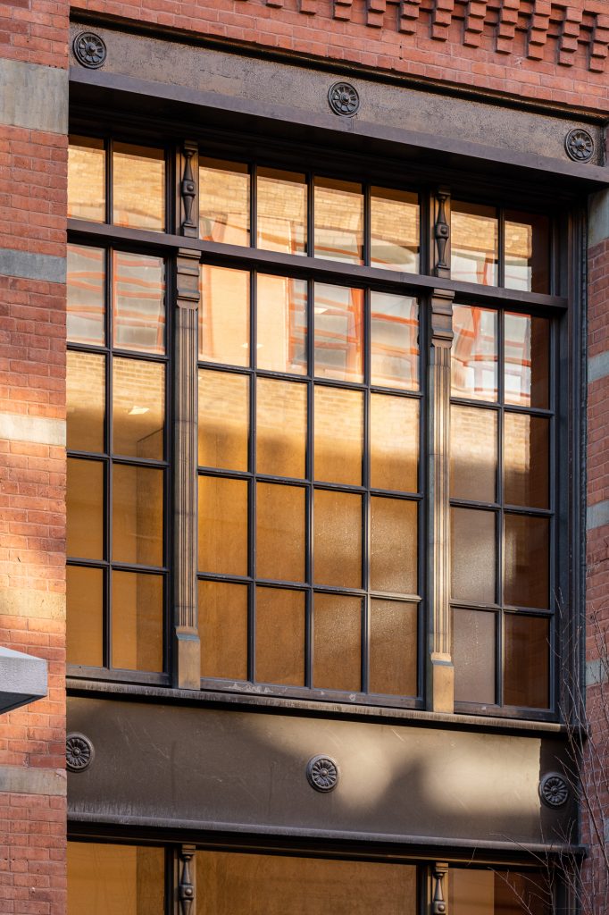 A large, multi-pane industrial-style window framed by red brick and concrete bands, featuring decorative black cast iron elements with floral rosettes and fluted pilasters, reflecting a neighboring building's façade in its glass, with warm light from the interior subtly visible behind the grid-like window structure, contributing to the architectural richness and historic character of the urban exterior.