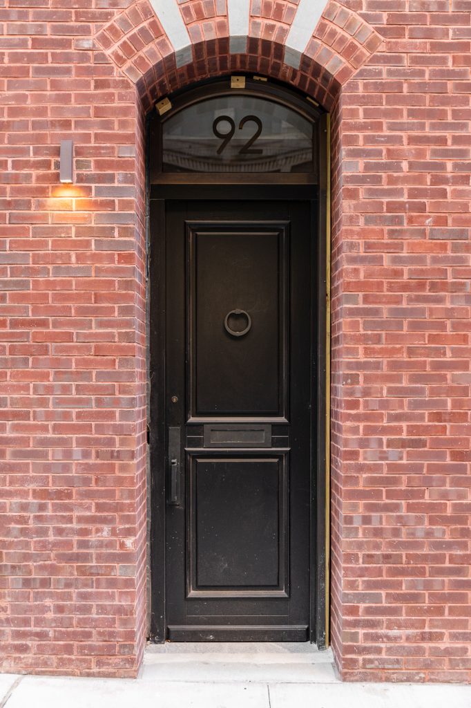 A black wooden front door with a circular knocker, a mail slot, and decorative recessed panels is set within a red brick building façade featuring a rounded archway made of alternating red and gray bricks, topped by a transom window with the number "92" in modern black numerals, all illuminated by a sleek, wall-mounted exterior light fixture on the left side of the doorframe.
