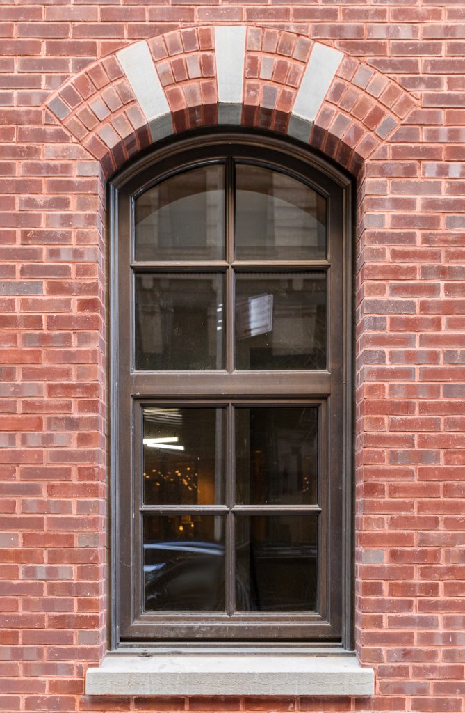 A tall arched window with a dark metal frame and six panes is symmetrically set into a red brick wall, topped by a decorative arch of alternating red and gray bricks, and featuring a gray stone sill at the base, while faint reflections of lights and surroundings are visible in the window glass.