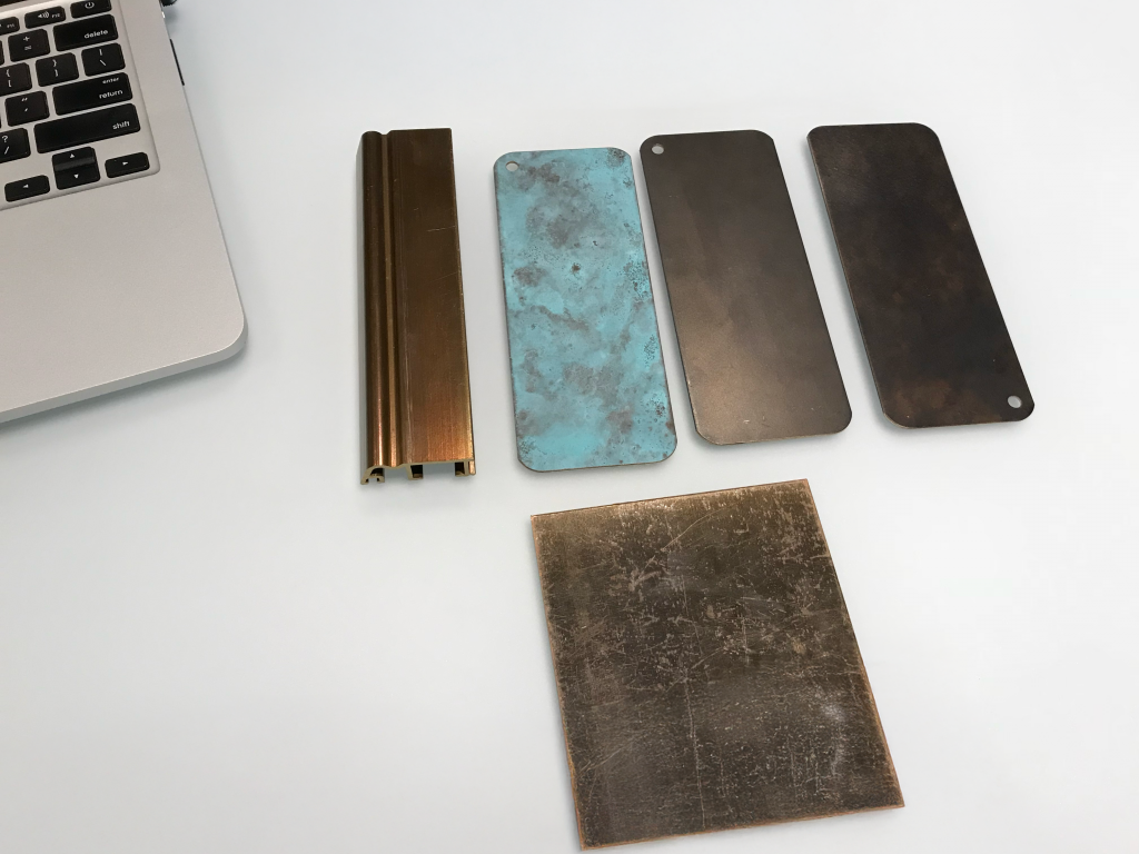 A collection of five metallic material samples arranged neatly on a white surface next to the bottom left corner of a silver laptop keyboard, including a bronze-toned extruded aluminum profile with dual channels, a patinated turquoise-colored copper sheet with mottled texture, two rectangular dark metal plates with rounded edges and small corner holes—one matte and the other with subtle discoloration—and a scratched, square-cut brass or bronze plate placed diagonally, all representing various metal finishes and oxidation states used in architectural or industrial design contexts.