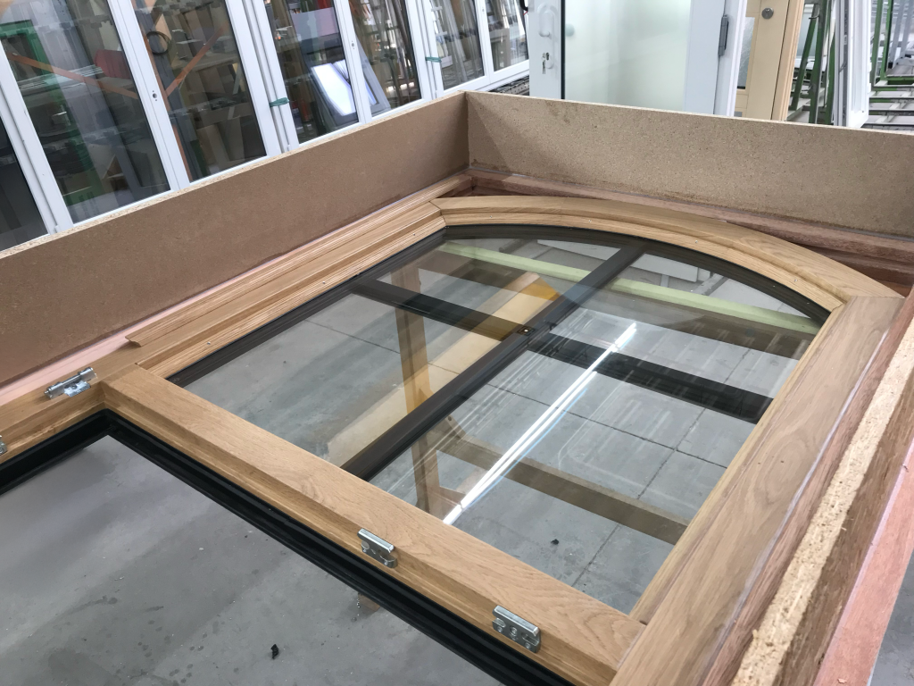 Large arched wooden-framed window unit with clear double-glazed panes and black muntin bars is positioned horizontally in a workshop setting, surrounded by MDF panels for protection, with visible hinges indicating an operable sash, while in the background, various other window and door assemblies are lined up vertically on racks in a well-lit industrial fabrication space.