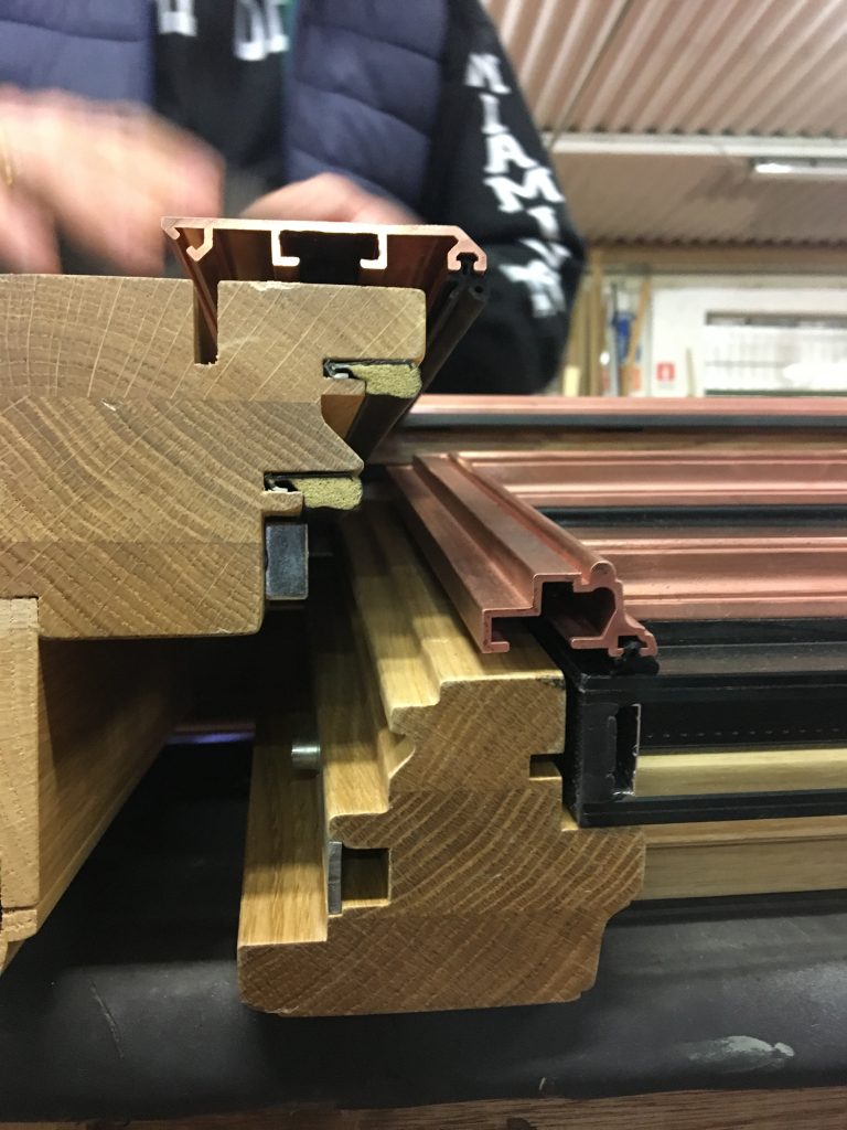 Close-up cross-sectional view of a high-quality wooden window frame featuring a complex joinery profile and multiple material layers, including a reddish-brown bronze cladding  fitted over the exterior for weather protection, precision-cut wood joinery elements forming insulation and sealing cavities, embedded gaskets and fasteners indicating a high-performance thermal or acoustic window system, with blurred hands of a person in the background suggesting manual assembly or inspection in a professional workshop setting.
