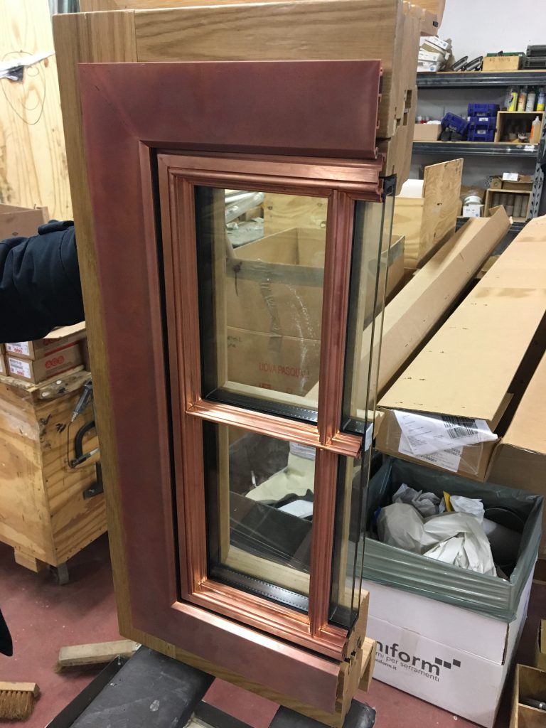 A meticulously crafted window mock-up featuring a rich copper cladding system with sharp, mitered corners and a warm, reddish metallic finish, integrated into a wooden frame with natural grain textures, displayed in a workshop setting with visible tools, cardboard boxes, wood components, and shelving in the background, highlighting precision fabrication and high-end material contrast for architectural applications.