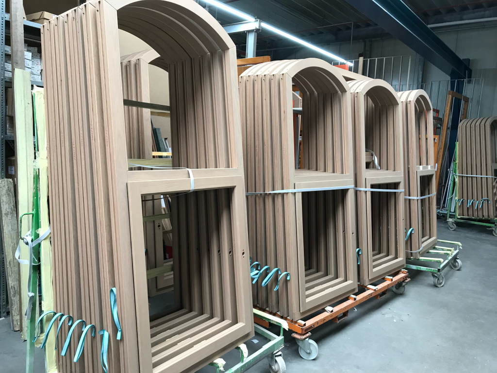 A series of meticulously crafted arched and rectangular wooden window frames uniformly stacked on wheeled carts in a spacious industrial workshop, each bundle neatly organized and secured with white straps and blue lifting loops, surrounded by structural steel beams, vertical storage racks, and overhead fluorescent lighting, reflecting a high level of precision manufacturing, large-scale production, and material handling in a professional carpentry or joinery facility.