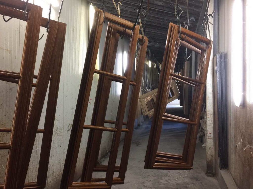 Multiple freshly varnished wooden window sashes with mullions are suspended vertically from metal hooks on an overhead conveyor system inside a narrow, industrial paint or drying booth, with fluorescent lights lining the walls and casting reflections on the glossy wood finishes, and the background receding into a dimly lit tunnel of hanging frames awaiting further processing.