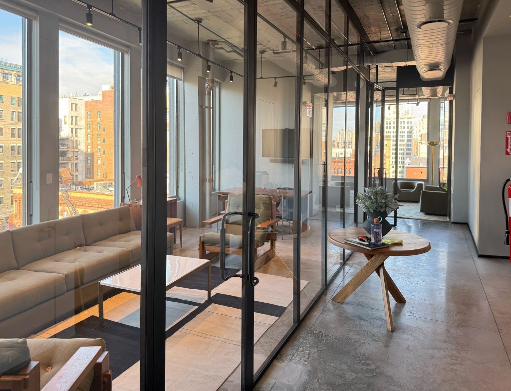 Modern office interior with floor-to-ceiling custom blackened steel partition walls dividing a lounge area furnished with beige tufted sofas, mid-century wooden armchairs, and a white rectangular coffee table, from a corridor lined with polished concrete floors and a round wooden table holding a plant, while large windows on the left flood the space with natural light and offer expansive views of a cityscape with varied architectural styles and colors, including brick and stone buildings under a clear blue sky.