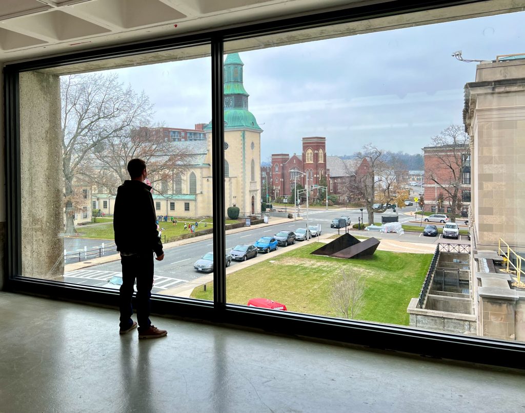Person standing in front of a massive floor-to-ceiling window inside a modern concrete building, gazing out at a picturesque urban landscape that includes a historic cream-colored church with a green copper steeple, a red-brick Gothic-style building, a sculpture on a manicured lawn below, parked cars lining the street, leafless trees indicating an early spring setting, and a river faintly visible in the distant background under an overcast sky.