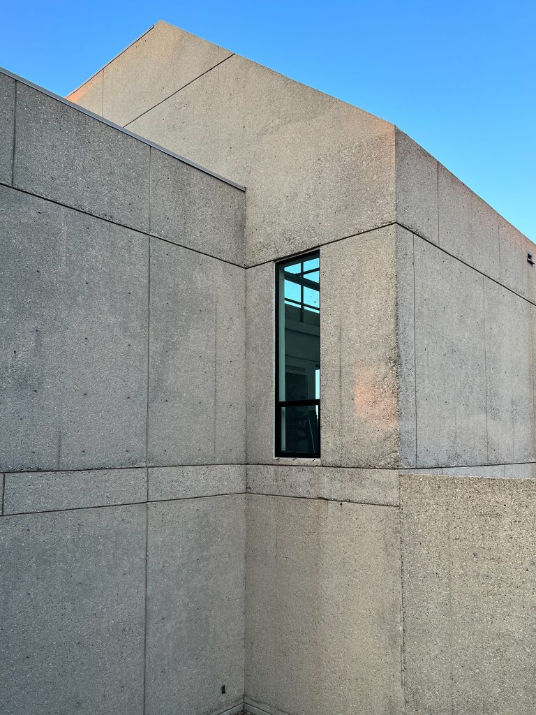 Concrete façade building with clean geometric lines and a prominent vertical window reflecting interior architectural elements, the raw textured concrete surfaces showing variations in color and weathering, set sharply against a clear blue sky, emphasizing the stark contrast between the structure’s solidity and the openness above.