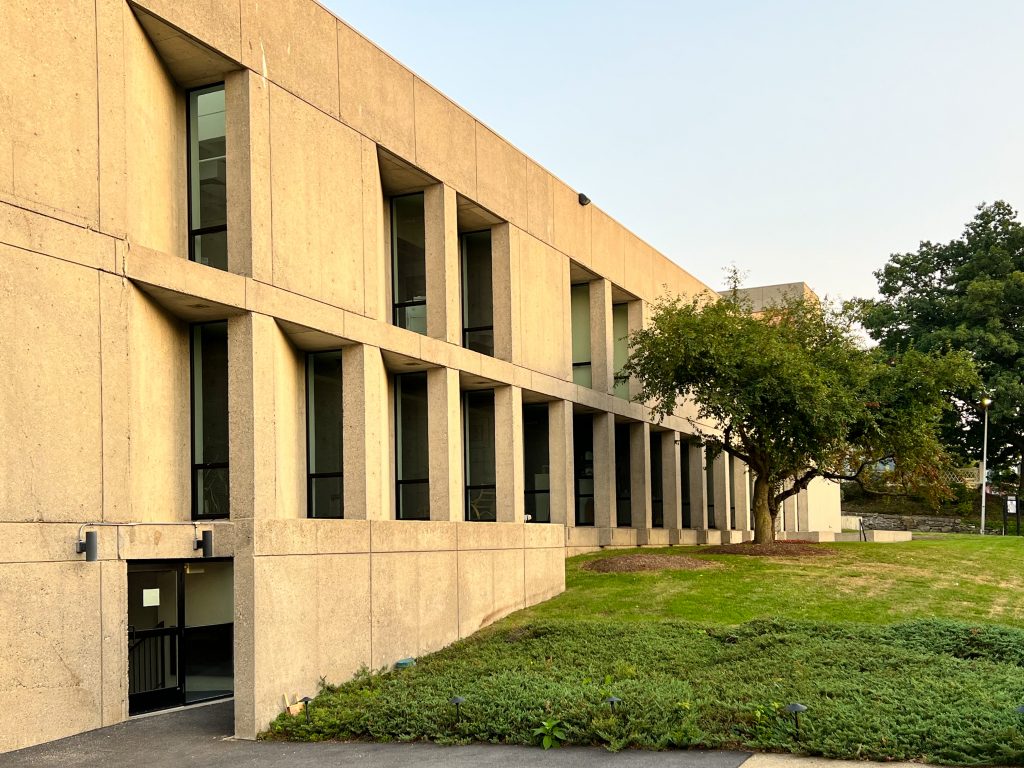 Modern concrete building featuring tall vertical windows recessed between massive structural columns, illuminated by the soft golden hue of late afternoon sunlight, with a well-maintained lawn and manicured shrubbery in the foreground, a lone tree providing shade near the center, and mature trees and a lamppost visible in the background, contributing to a serene and orderly architectural landscape.