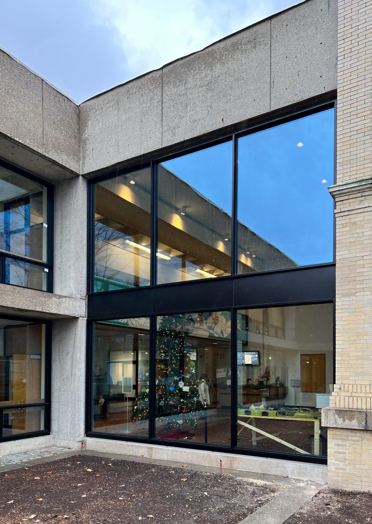 Corner view of a concrete building with expansive black-framed windows revealing a warmly lit interior decorated for the holidays, including a Christmas tree adorned with ornaments and lights, positioned beside a light yellow brick structure that provides a stark material contrast, all under a dusky sky that reflects subtly in the tall glass panes.