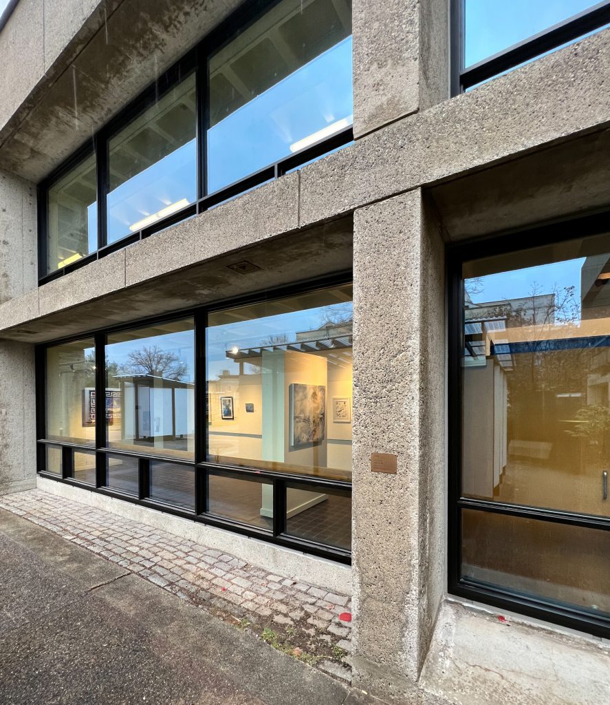 Concrete and glass façade of a two-story building features large black-framed windows revealing a brightly lit interior art gallery with several paintings and artworks on white and pale blue walls, while the exterior shows a rain-dampened cobblestone and asphalt walkway under a gray-blue sky with bare trees reflected in the glass, emphasizing the clean lines, industrial textures, and transparency between indoor exhibition space and outdoor urban environment.