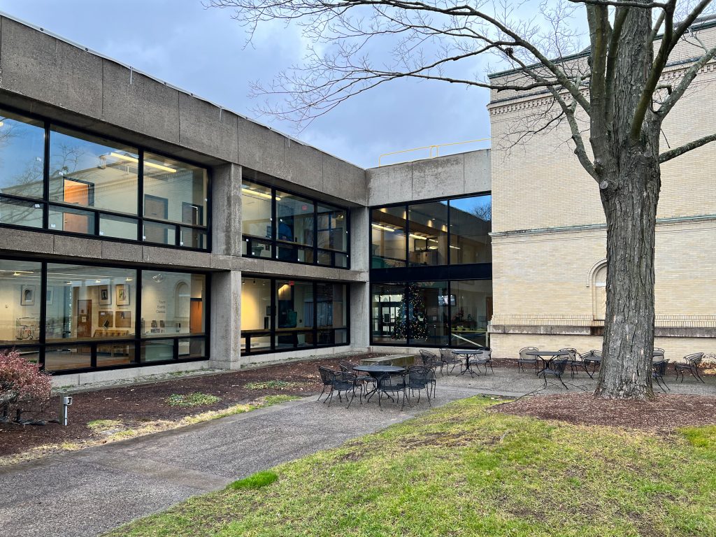 Concrete building with two stories of large black-framed windows revealing interior spaces filled with art displays and a Christmas tree, sits adjacent to a yellow-brick historic structure with classical details, while in the foreground a leafless tree stands near a paved courtyard with empty metal patio tables and chairs, surrounded by early spring dormant landscaping, under a moody blue-gray sky.