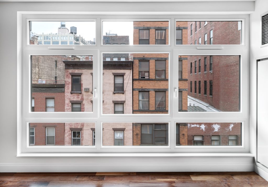 Large rectangular window with white framing dominates the wall of a minimalist interior space with glossy wood flooring, revealing a close-up view of neighboring brick and stone buildings with varied textures, window styles, and faded facades, including red, tan, and rust-colored exteriors, creating a dense, layered urban environment that emphasizes architectural repetition and the compressed perspective of city living.