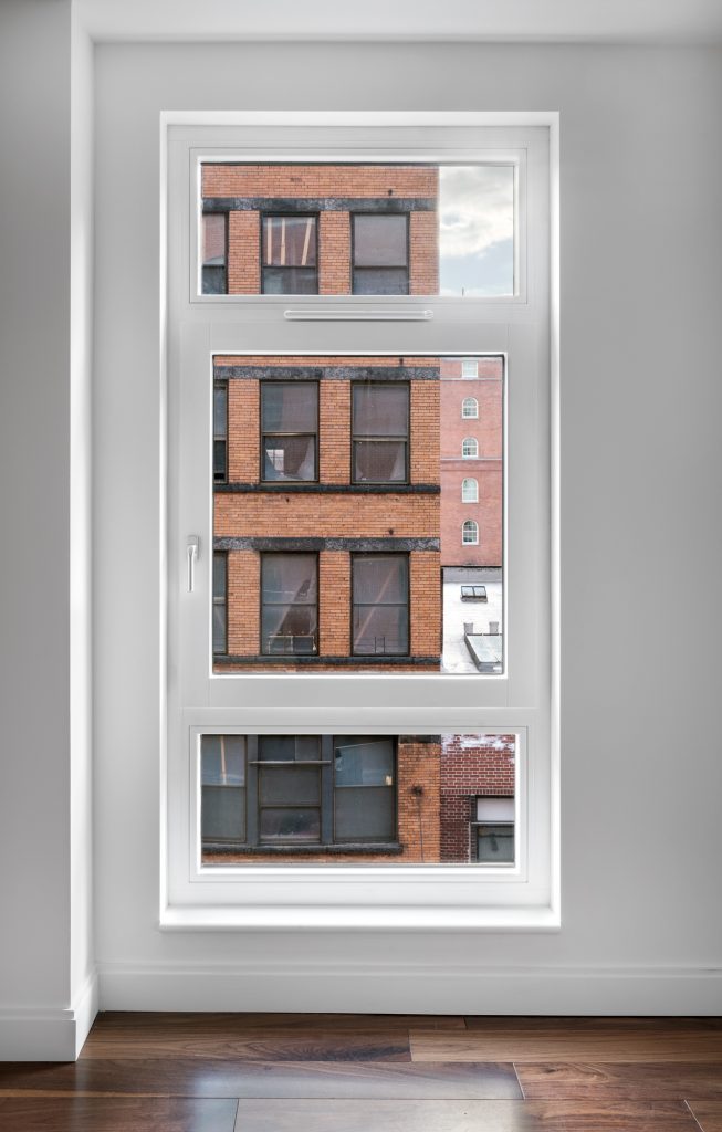 Tall, narrow modern window with crisp white framing set into a clean white wall and overlooking a dense cityscape, where closely packed mid-rise buildings feature red and orange brick facades, dark-framed industrial windows, and worn architectural details, all contrasted against the soft natural light reflecting on polished wood flooring inside the minimalist interior space.