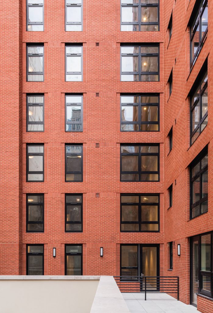 Narrow urban courtyard view framed by red brick walls on three sides, featuring an array of black-framed rectangular windows in a repetitive grid pattern, a flat light-colored patio area at the bottom with a metal handrail and wall-mounted lights, and glass double doors serving as an entryway into the building, with reflections of neighboring structures subtly visible in the windows.