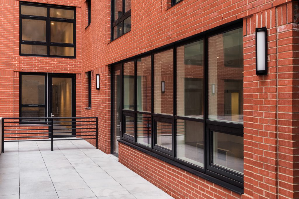 Ground-level exterior view of a modern building façade combining smooth gray concrete panels with red brickwork, centered around a large black-framed glass window and door assembly that reflects the surrounding urban streetscape, revealing an inviting interior with dim warm lighting, visible lounge chairs, a coffee table with a small floral centerpiece, and a unique smaller operable window inset above the main glass panels, flanked by a minimal vertical wall sconce on the left side.