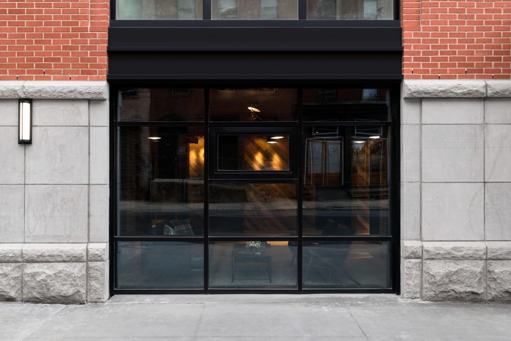 Ground-level exterior view of a modern building façade combining smooth gray concrete panels with red brickwork, centered around a large black-framed glass window and door assembly that reflects the surrounding urban streetscape, revealing an inviting interior with dim warm lighting, visible lounge chairs, a coffee table with a small floral centerpiece, and a unique smaller operable window inset above the main glass panels, flanked by a minimal vertical wall sconce on the left side.