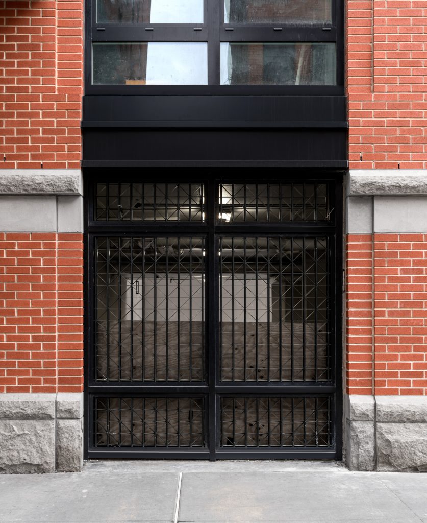 Street-level view of a secured black metal-framed entryway with crisscross lattice-style security grilles covering large glass panels, set within a red brick and gray stone-clad building façade, featuring a solid concrete sidewalk in the foreground, visible fluorescent lighting inside the partially finished interior space, and a black architectural band separating the entry from upper-level residential or commercial windows.