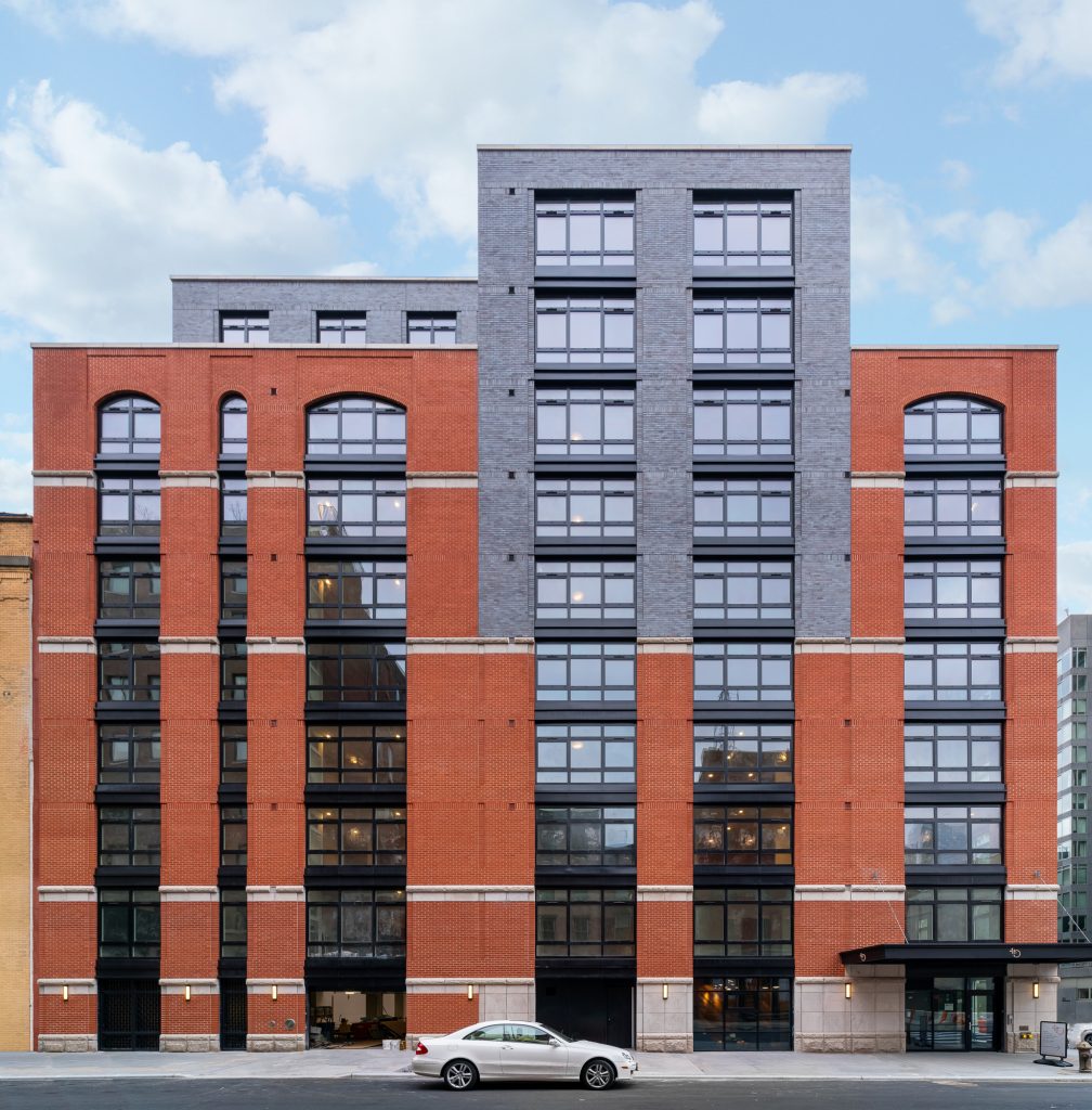 Symmetrical multi-story urban building featuring a blend of modern and traditional design elements, with a red brick facade accented by gray brick on the central section, large industrial-style black-framed windows in uniform rows across all levels, arched windows near the top for visual interest, a street-level entrance canopy to the right, and a white sedan parked along the sidewalk in front of the structure under a bright, partly cloudy sky.
