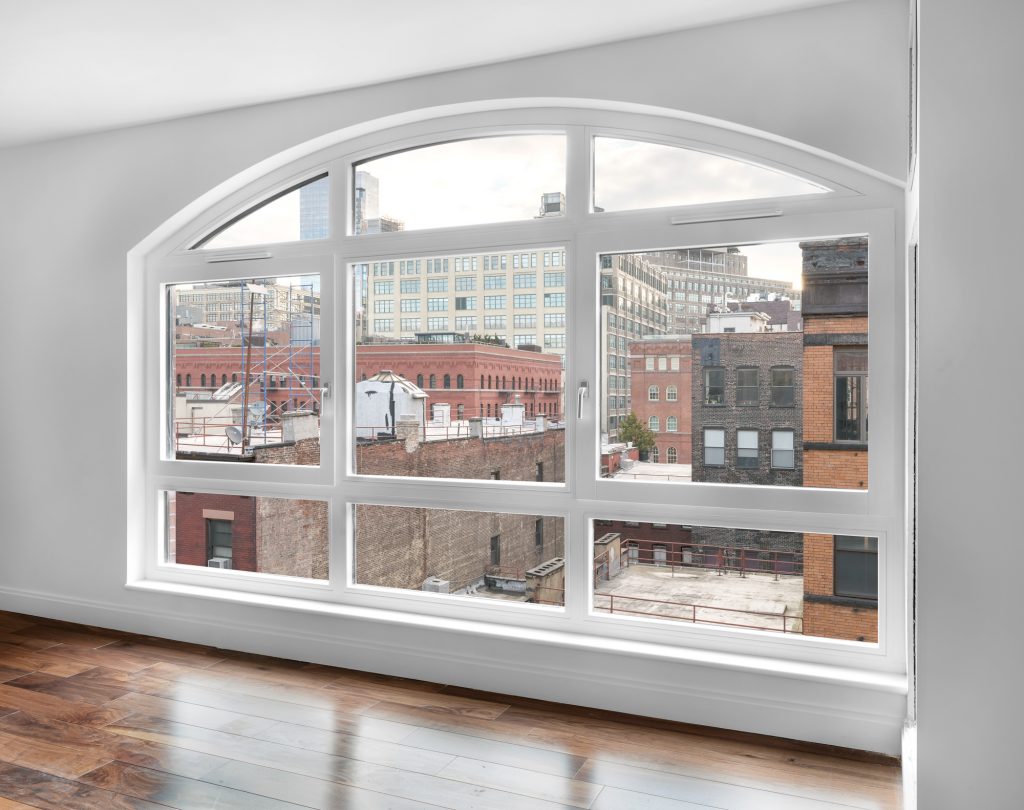 Large arched white-framed window with multiple panes fills the wall of a bright modern interior with glossy wooden floors, offering a panoramic view of a densely packed urban streetscape composed of historic brick buildings, industrial rooftops with mechanical equipment and ladders, and taller glass-and-steel commercial structures in the background, all under a partly cloudy sky that adds depth and softness to the geometric rhythm of the cityscape.