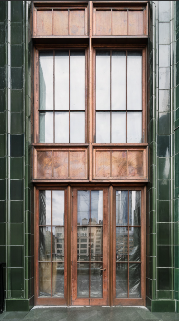 Tall, elegant façade featuring three vertically stacked sections of large, gridded windows and doors framed in warm, weathered copper metal, set within a deep green, glossy-tiled exterior wall, reflecting the surrounding urban architecture in the glass panels, with the bottom section composed of double doors opening onto a terrace or balcony, flanked by pilasters.