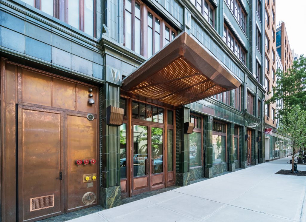 Street-level view of a sophisticated building entrance featuring a bronze-toned canopy with perforated underlighting, warm wooden-framed glass double doors, surrounding green-patinated metal and polished dark stone facade, adjacent to a copper utility panel with red and yellow valve fixtures, and flanked by tall street-facing windows that reflect the urban streetscape, all set along a clean, wide sidewalk lined with young trees.