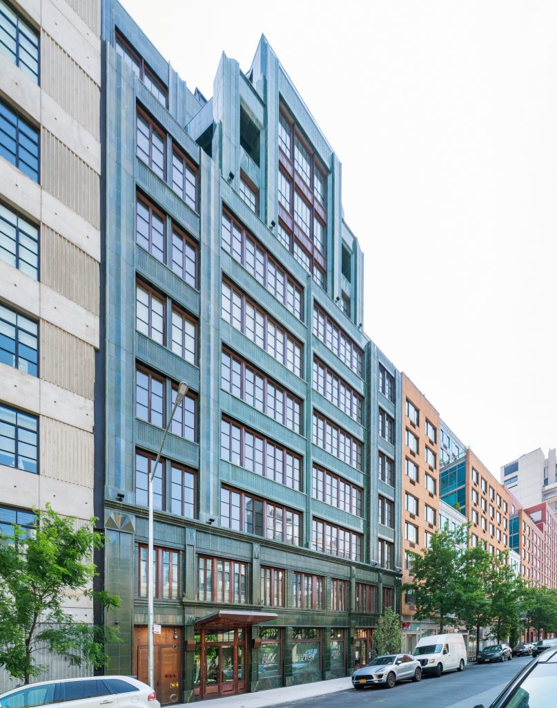 Tall mid-rise building with a distinctive green-patinated facade and large grid-style windows framed in dark wood, featuring strong vertical architectural lines and stepped upper levels that create an profile, positioned along a clean city street lined with parked cars and mature trees, flanked by neighboring structures in contrasting beige concrete and warm-toned brick, under a bright overcast sky that diffuses light evenly across the urban scene.
