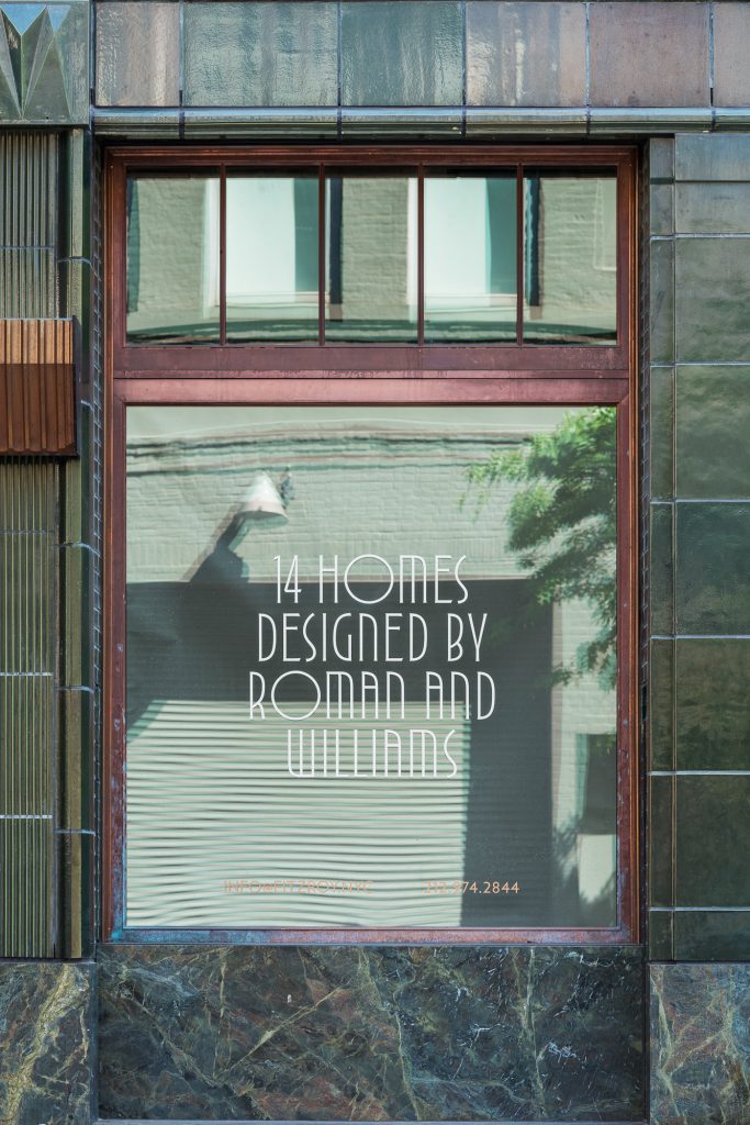 Close-up of a large street-level window framed in dark reddish-brown wood set within a green-patinated metal and dark marble facade, reflecting the opposing building’s pale brick wall, windows, and outdoor light fixture, with bold white text reading “14 Homes Designed by Roman and Williams” along with smaller contact details near the bottom, partially shadowed by nearby trees that cast organic shapes onto the glass.