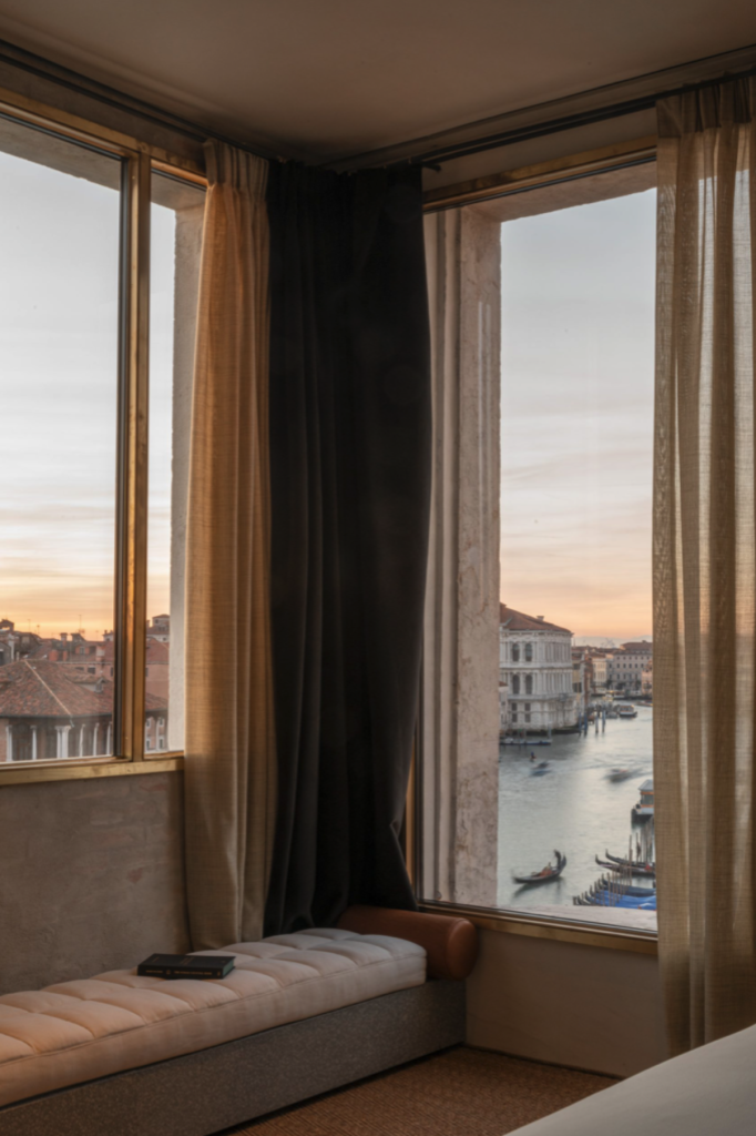 In a contemporary guest room at The Venice Venice Hotel, two expansive, satin brass-framed Secco Sistemi OS2 windows meet at a corner to reveal a panoramic, golden-hour view of the Grand Canal, where gondolas glide past the historic 13th-century Palazzo Ca' da Mosto, while the interior features a minimalist, tufted cream-colored bench, a solitary book, and a sophisticated combination of sheer linen and heavy dark floor-to-ceiling drapes.