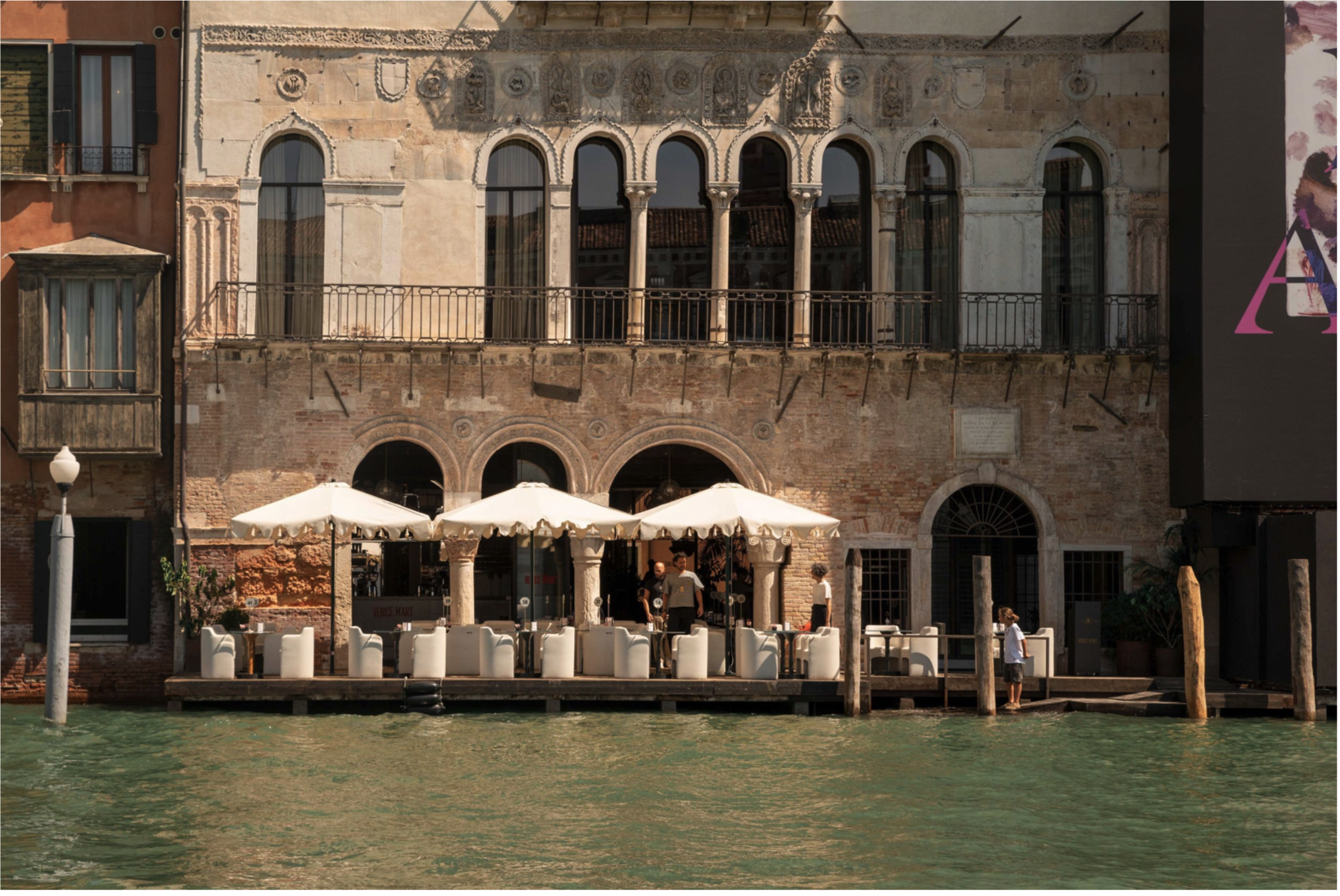 A sun-drenched view from the Grand Canal captures the historic 13th-century Venetian-Byzantine facade of Ca' da Mosto, now home to The Venice Venice Hotel, featuring an elegant upper-level loggia with slender columns and six stilted arches, a terrace with white umbrellas and minimalist seating on the ground floor, and weathered brick walls adorned with faded decorative patere and marble coats of arms.