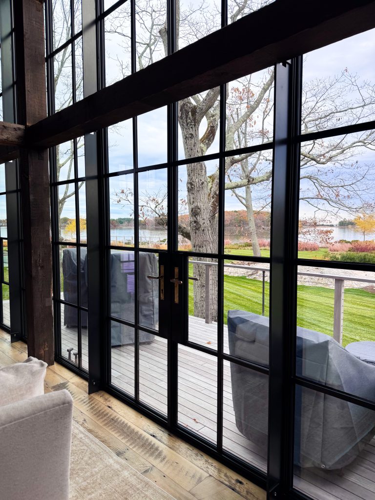Interior view of a modern rustic living space featuring tall black steel–framed glass doors with a grid pattern and slim mullions, a dark structural timber beam crossing overhead, wide natural wood plank flooring with visible grain and wear, and a seamless visual connection to an outdoor wooden deck with covered seating, leafless trees, manicured green lawn, and a calm waterfront landscape under an overcast sky, emphasizing transparency, material contrast, and indoor–outdoor continuity.