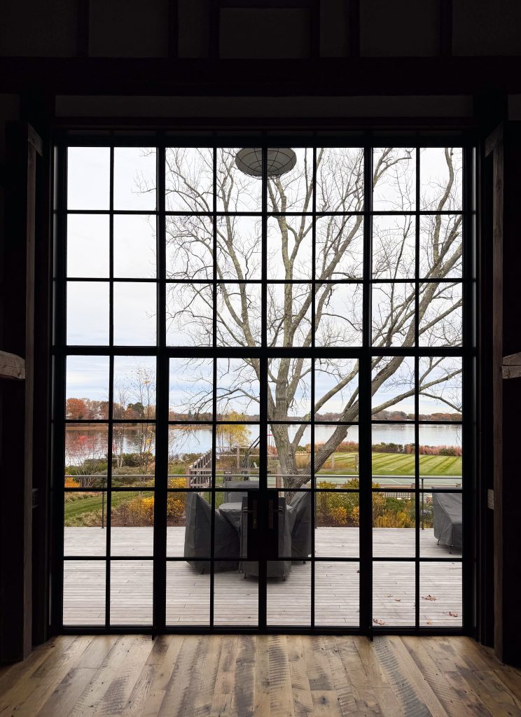 An interior view through a monumental black steel-framed window wall with integrated double doors reveals a seamless transition to an expansive light-wood deck, where covered patio furniture sits before a starkly elegant bare tree, all framed against a serene lakeside landscape featuring a manicured lawn, autumn-colored foliage, and a calm body of water under a soft, overcast sky.