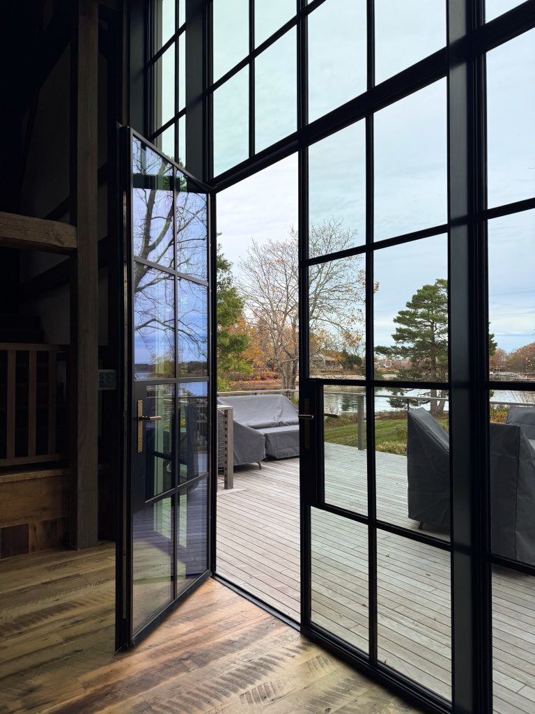 An interior view through a monumental black steel-framed window wall with an open swinging door reveals a seamless transition from rustic wide-plank hardwood floors to an expansive wooden deck, where grey-covered outdoor furniture sits before a serene lakeside landscape featuring a manicured lawn, autumn-colored trees, and a calm body of water under a soft, overcast sky.