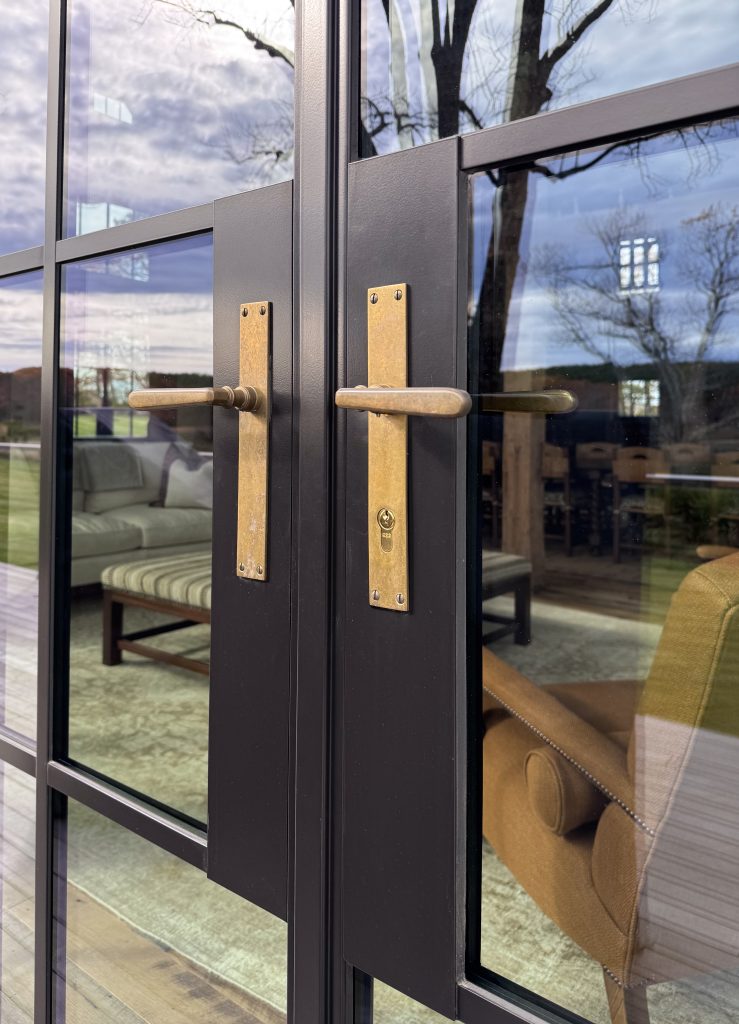 Close-up view of modern black-framed glass double doors with tall rectangular brass backplates and matching horizontal brass handles, reflecting leafless trees and a cloudy sky outside, while revealing through the glass a cozy interior with a light-colored sofa, patterned bench, wooden chairs, textured rug, and a brown upholstered armchair, emphasizing a blend of contemporary architecture, natural light, and warm interior design details.