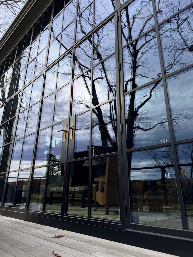A low-angle shot captures the facade of a modern building featuring floor-to-ceiling windows in black steel frames, centered by double swinging doors with elegant brass handles that reflect the silhouettes of bare trees and a dramatic, cloudy sky, while the transparent panels reveal a cozy interior with an armchair and sofa situated on a light-colored wood-plank terrace.