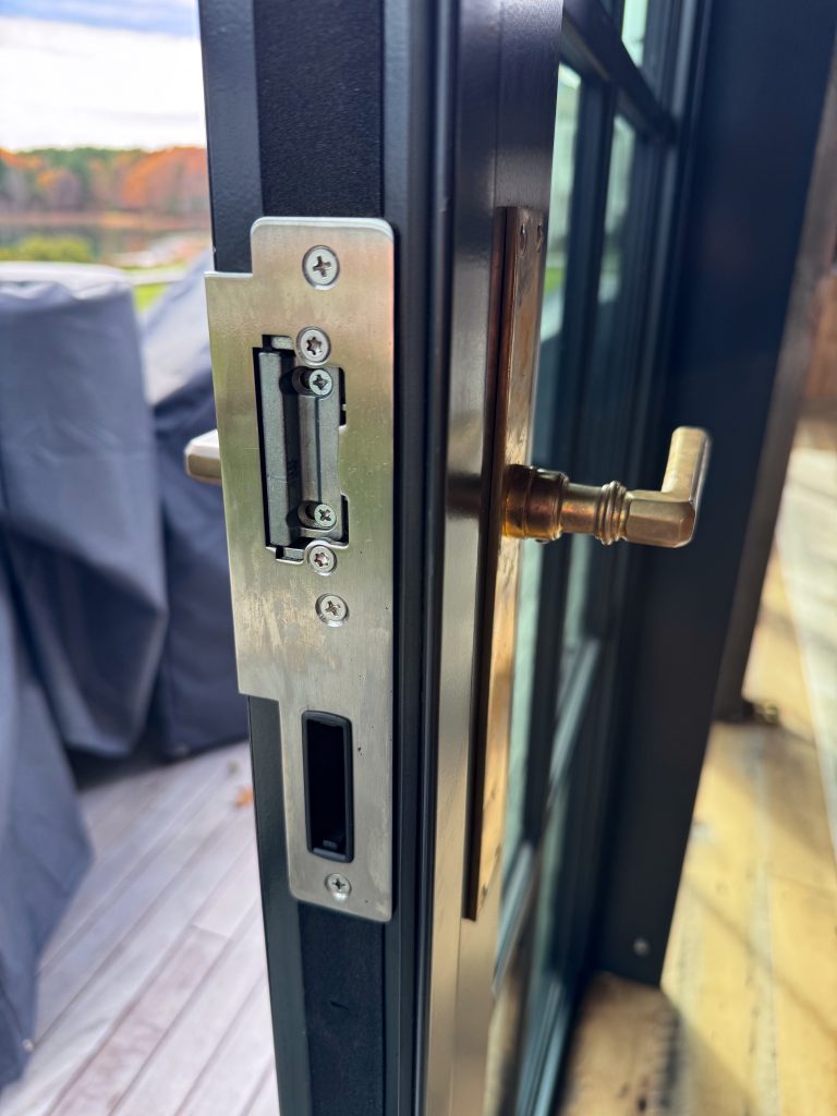 A close-up, high-angle shot of a modern black sliding door frame features an intricate stainless steel mortise lock assembly with exposed mounting screws and a rectangular latch mechanism, while a solid brass lever handle with a tiered design extends from a matching backplate, all set against a blurred background of an outdoor deck with grey patio furniture covers and a scenic view of autumn-colored trees and water under a bright sky.