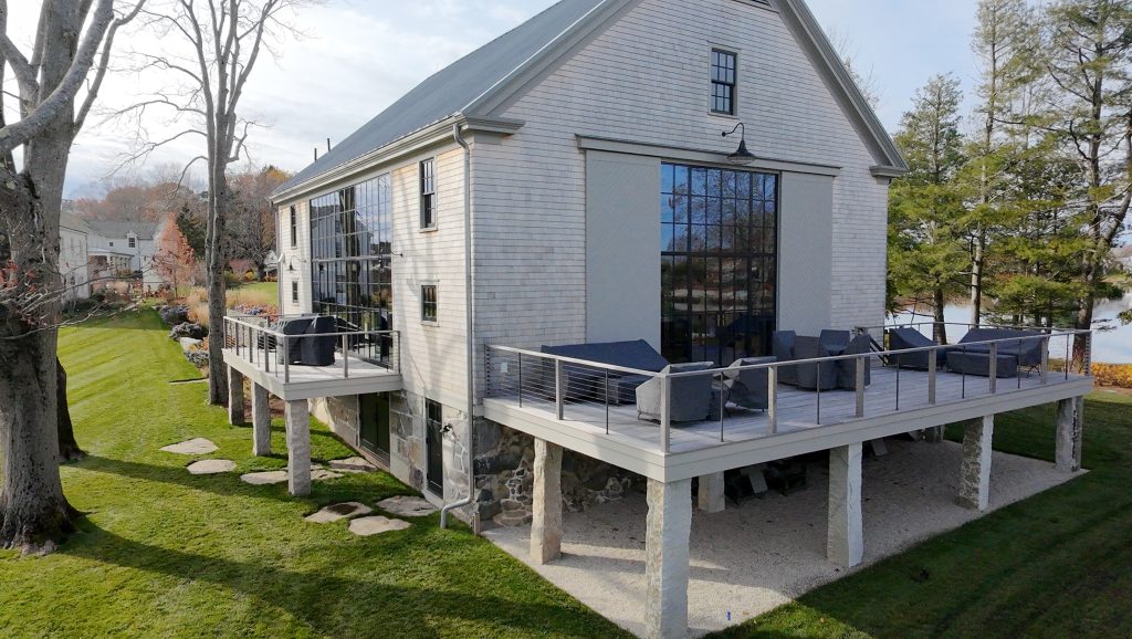 Exterior view of a renovated barn-style house with light gray shingle siding and a steep metal gabled roof, featuring large black steel-framed grid windows and glass doors that reflect surrounding trees, elevated wraparound decks with cable railing and outdoor lounge furniture supported by stone and concrete piers, a stone foundation at ground level, manicured green lawn with stepping stones and mature leafless trees, and a tranquil waterfront landscape in the background under soft daylight, highlighting modern coastal architecture blended with rustic materials and indoor-outdoor living